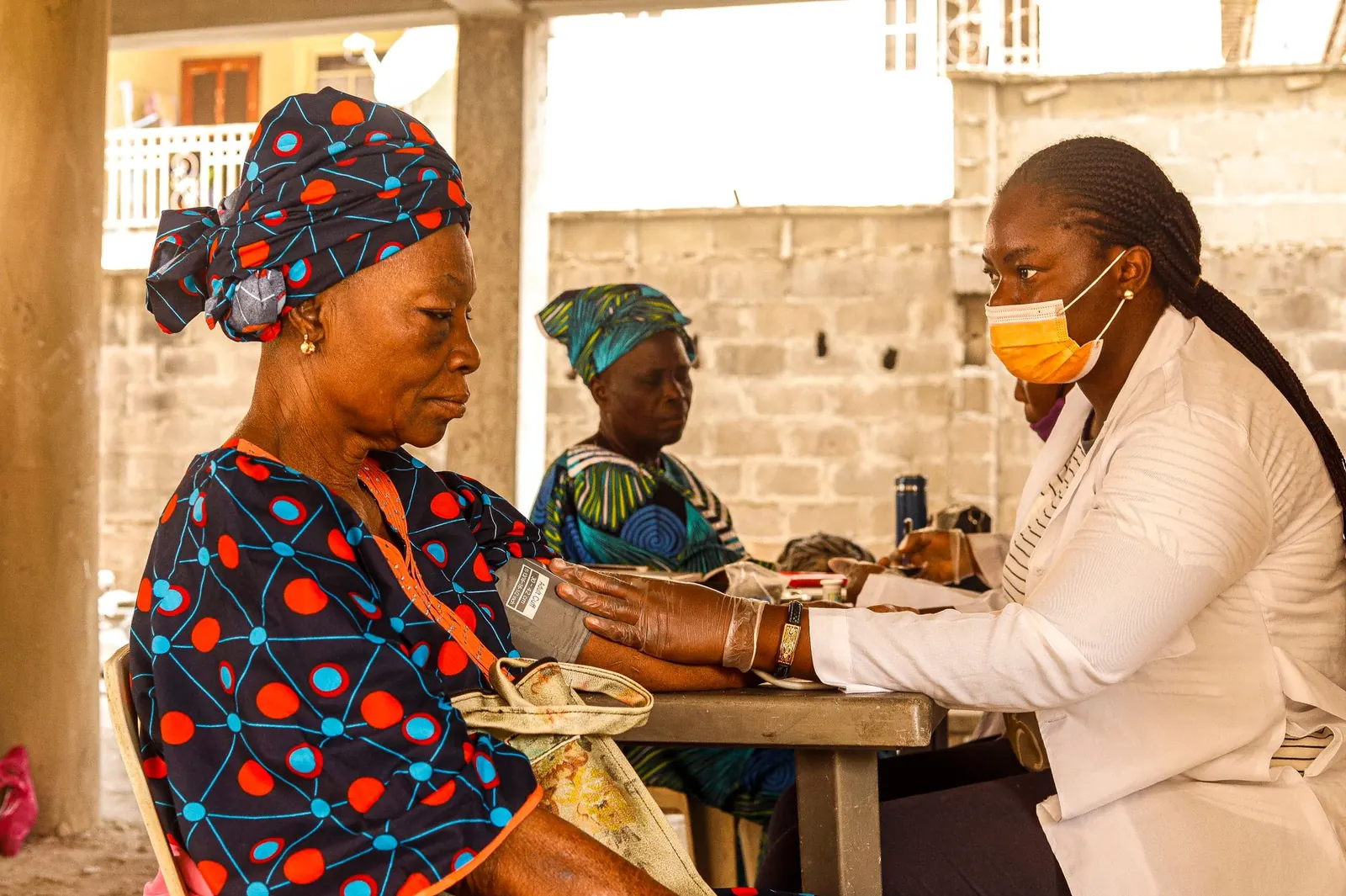 Health worker in a white coat checking an older woman's blood pressure at an outdoor screening