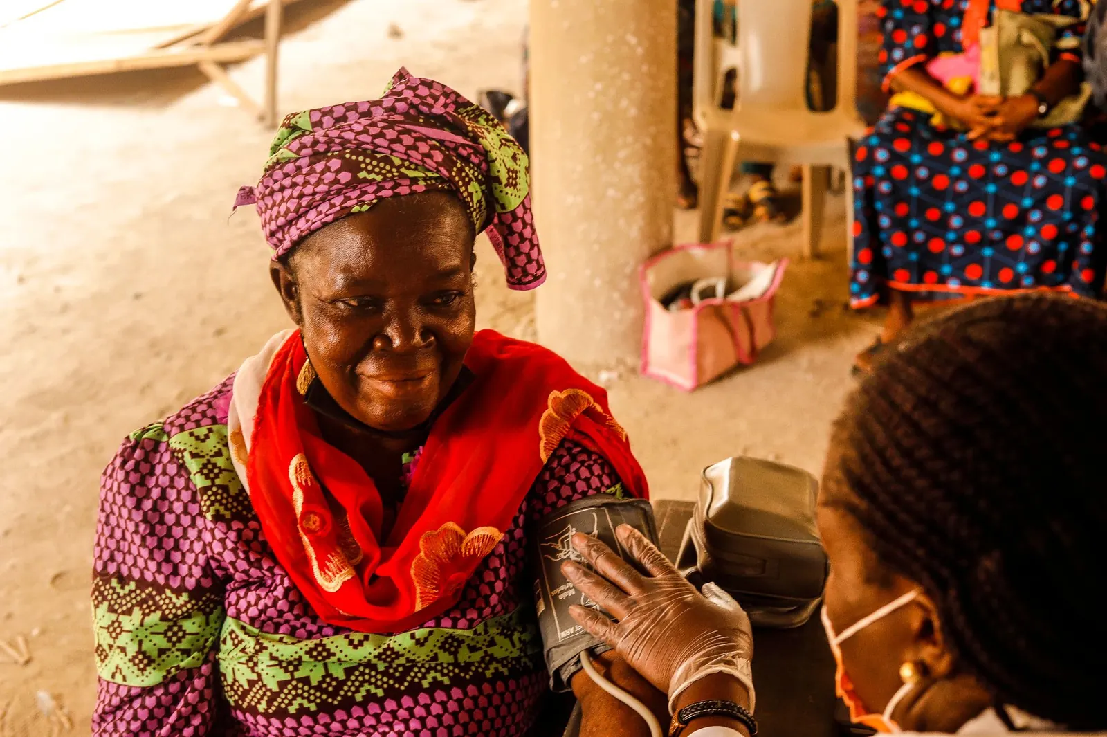 Smiling woman in a headwrap and patterned dress having her blood pressure checked at a community screening